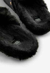 Svart tofflor med pälsfoder som har en mjuk och plyschig inredning samt ett slätt yttermaterial med en diskret logotag.