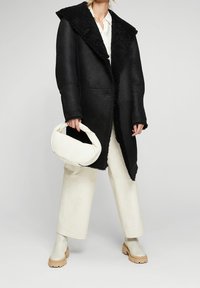 Mujer vestida con un abrigo de shearling negro, pantalones de pierna ancha blancos y botas de tobillo crema, sosteniendo un bolso blanco con asas retorcidas.