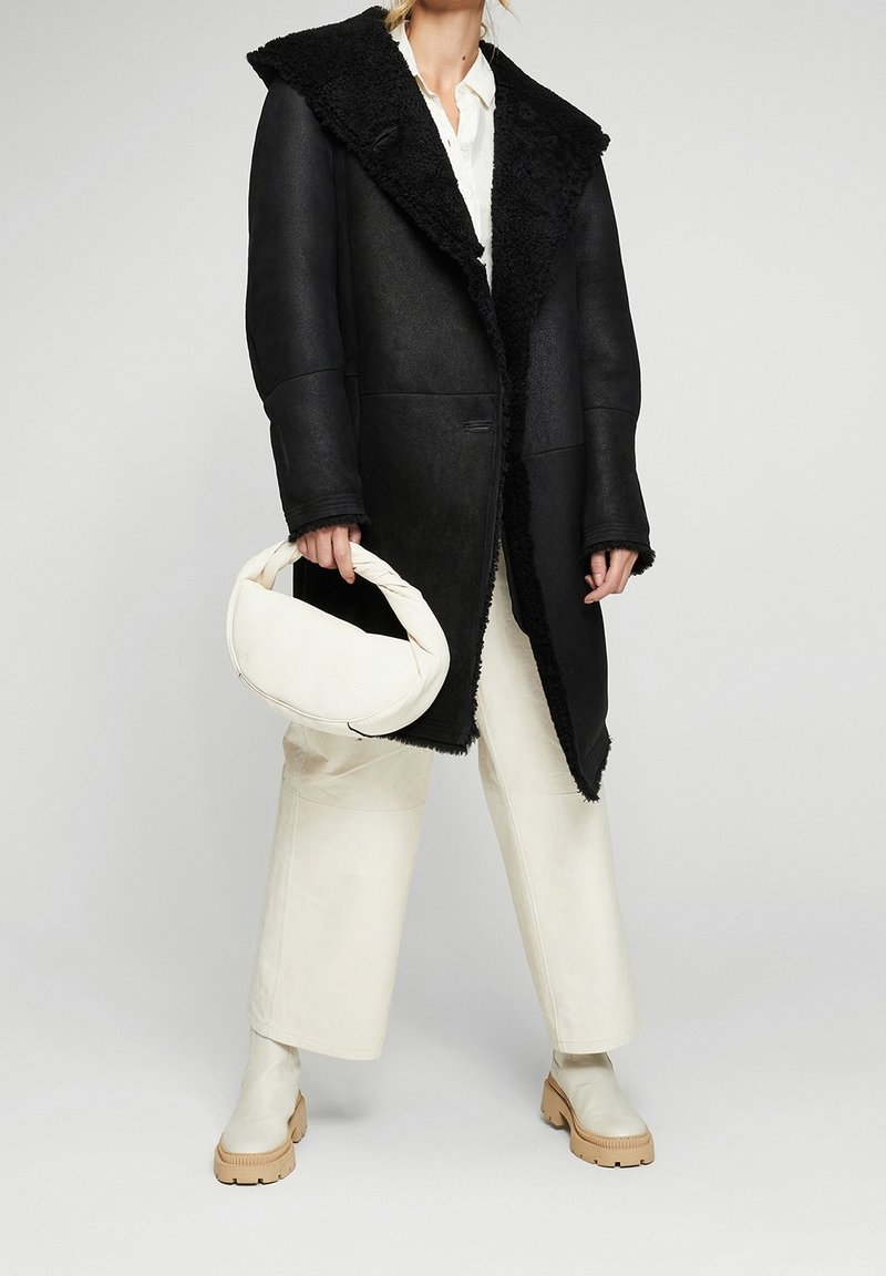 Mujer vestida con un abrigo de shearling negro, pantalones de pierna ancha blancos y botas de tobillo crema, sosteniendo un bolso blanco con asas retorcidas.