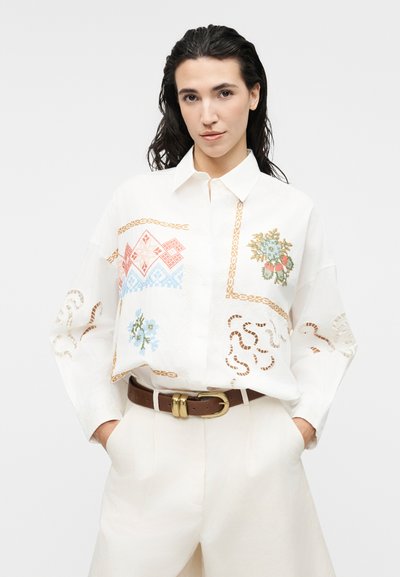 Γυναίκα με σκούρα μαλλιά που φοράει λευκό πουκάμισο με πολύχρωμη γεωμετρική και ανθική κεντητική, λευκό παντελόνι και καφέ ζώνη.