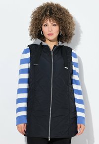 Chaleco acolchado negro con cierre de cremallera y capucha, combinado con una camiseta de manga larga a rayas en azul y blanco. Tejido texturizado con detalles de cordón.