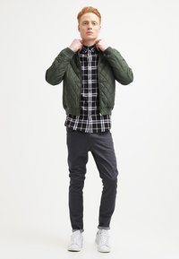 Jachetă bomber verde matlasată peste o cămașă în carouri alb-negru, asortată cu pantaloni gri închis și adidași albi.