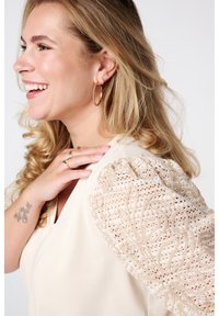 Beige blouse met textuurkant mouwen met geborduurde patronen, v-hals ontwerp en een vloeiende pasvorm. Geaccessorieerd met ringoorbellen.