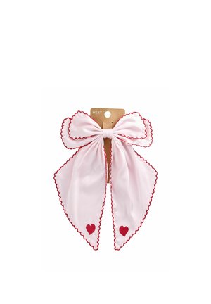 Große blassrosa Stoffschleife mit roten gezackten Kanten und roten Herz-Akzenten an den Enden, befestigt an einer Pappe mit der Aufschrift "NEXT."