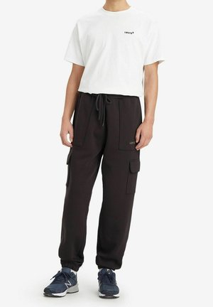 Person trägt ein weißes Levi's T-Shirt, schwarze Cargo-Jogginghosen mit Taschen und blaue New Balance Sneakers, steht vor weißem Hintergrund.