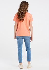 Perzikkleurig katoenen t-shirt met korte mouwen, een relaxte pasvorm en een ronde zoom, gecombineerd met lichtblauwe spijkerbroeken en witte sneakers.