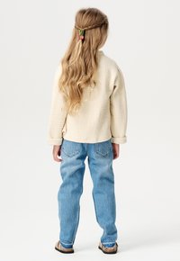 Hellbeige gesteppte Jacke mit umgeschlagenen Ärmeln, blaue lässige Jeans und braune Sandalen. Haar mit bunten Clips gebunden, wellig herabfallend.