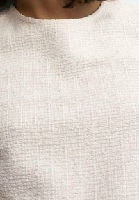 Nainen, joka on pukeutunut valkoiseen teksturoituun yläosaan, jossa on pyöreä pääntie, paljastaen kaulan ja ylärinnan alueen.