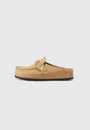 Beige Clogs mit einem breiten Riemen und goldener Schnalle, aus weichem Material gefertigt, mit einer Kork-Zwischensohle und einer strukturierten schwarzen Gummisohle.
