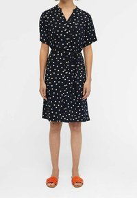 Robe marine à pois blancs, à manches courtes, décolleté en V, taille cintrée, longueur genoux. Portée avec des sandales à glisser orange.