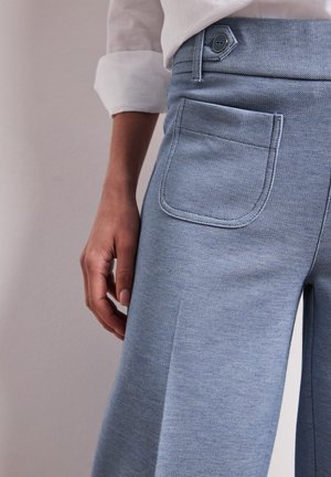 Persona che indossa pantaloni azzurri strutturati con tasca applicata frontale e una camicia bianca a maniche lunghe, mano rilassata al lato.