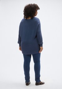 Μπλε σκούρο ριμπ πουλόβερ με άνετη γραμμή, μακριά μανίκια και καμπυλωτή βάση, συνδυασμένο με στενά σκουρό μπλε τζιν και καφέ επίπεδα παπούτσια.