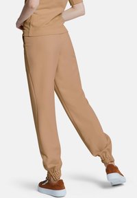 Beige taps sweatpants met elastische boorden, gemaakt van een zachte stof. Heeft zijzakken en een gladde textuur. G gedragen met bruine sneakers.