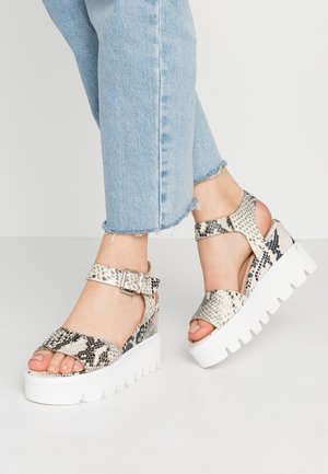 Feet wearing light blue cropped jeans and white platform sandals with black and grey snake print straps and buckles.