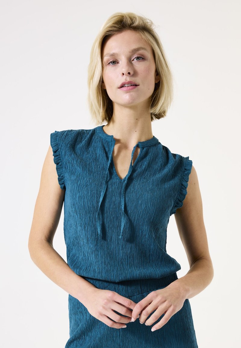 Teal top met een gestructureerde afwerking, korte geruffelde mouwen en een keyhole halslijn. Zachte stof met een ontspannen pasvorm, gedragen met bijpassende kleding.