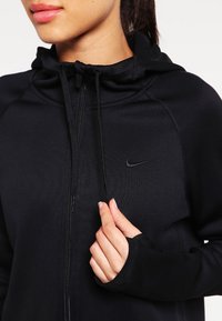Černá Nike mikina na zip s přiléhavým střihem, klokankovou kapsou, nastavitelnou kapucí a malým logem na hrudi. Hladká textura.