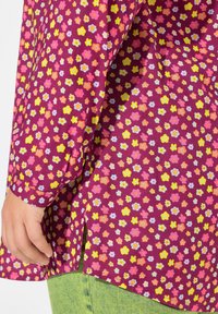 Lilla bluse med lange ærmer, prydet med flerfarvede blomster i pink, gult og blåt. Indeholder sidesplitter ved kanten.