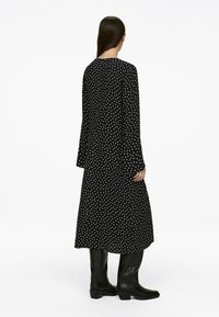 Schwarzes langärmliges Kleid mit weißen Punkten, fließendes Design, bis zur Mitte der Waden reichend, kombiniert mit knielangen schwarzen Stiefeln. Glatte Textur.