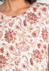 Top blanco con intrincados patrones florales y de paisley en rojo-borgoña. Tela suave con escote redondo y un sutil detalle de textura.