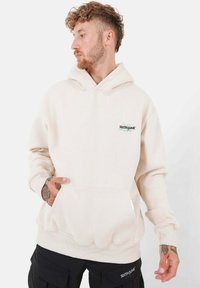 Beige Kapuzenpullover aus weichem Stoff mit einer Fronttasche, gerippten Bündchen und Saum sowie einem kleinen Logo auf der Brust.