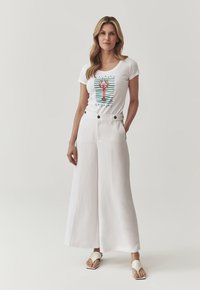 Weißes T-Shirt mit Hummergrafik auf blau-weißen Streifen, kombiniert mit hochtaillierten, weit geschnittenen weißen Hosen und neutralen Sandalen.
