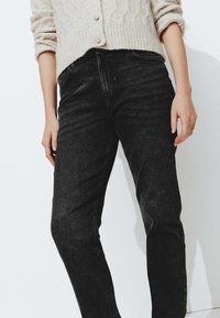 Jean en denim noir avec une coupe droite, affichant une légère décoloration, un design à cinq poches et une taille mi-haute. Un pull en maille neutre au-dessus.