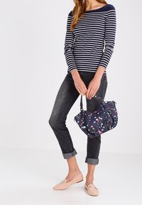 Laavupussissa on värikkäitä pilkkuja, useita lokeroita ja kultasävytteistä metalliosaa. Mallilla on päällä raidallinen paita, harmaat farkut ja vaaleat kengät.