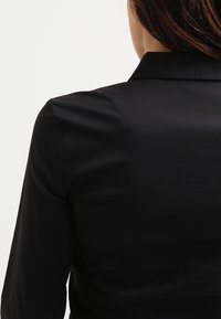 Czarna koszula o gładkiej, błyszczącej fakturze, z wyrazistym kołnierzykiem i dopasowanym krojem. Tył jest gładki, bez dodatkowych detali.