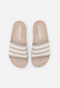 Šľapky Adidas so syntetickým vrchom v béžovej farbe, tromi bielymi pruhmi, textúrovanou vnútornou stielkou a zaoblenými okrajmi.