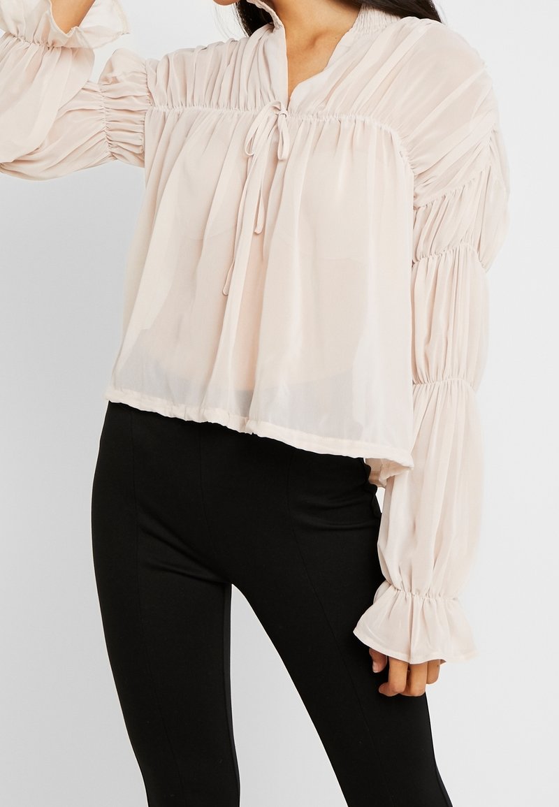 Femme portant un chemisier ample, transparent, rose pâle avec des manches longues froncées et un pantalon noir ajusté sur un fond uni.