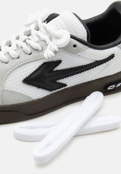 Λευκό αθλητικό παπούτσι με υφή, μαύρο λογότυπο βέλους, καφέ σόλα και λευκή στερέωση κορδονιών, συνοδευόμενο από δύο επιπλέον λευκά κορδόνια.