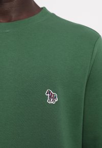 Chemise vert foncé avec un petit logo zèbre coloré brodé sur le côté gauche de la poitrine, portée par une personne à la peau foncée.