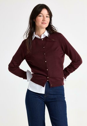 Kvinde med langt mørkt hår iført en hvid skjorte med krave under en knappet bordeaux cardigan og blå jeans, stående med hænderne på hofterne.