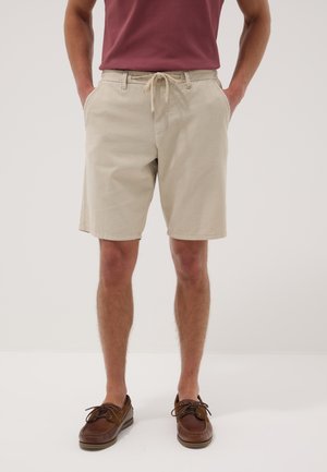 Uomo con pantaloncini beige con coulisse, scarpe da barca in pelle marrone e t-shirt bordeaux, in piedi con le mani nelle tasche.