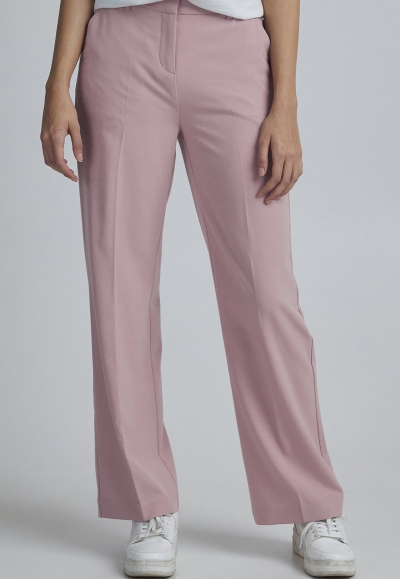 Personne portant un pantalon droit rose clair et des baskets blanches, debout devant un fond gris clair uni.