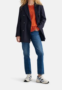 Manteau bleu marine croisé, pull orange à torsades, jeans bleu clair et baskets blanches avec des accents bleus.