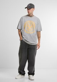Grå oversized t-skjorte med gul grafisk trykk og tekst, kombinert med svarte løse jeans og hvite sneakers. Grå caps båret på skrå.