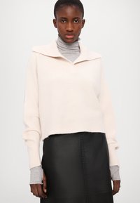 Ljust rosa ribbad tröja med bred krage, bärs över en grå polotröja, ihop med en svart läderskjorta med texturerade accenter.