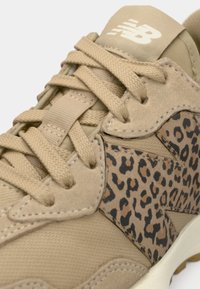 Gros plan d'une basket beige avec des lacets épais et un détail imprimé léopard sur la partie latérale, avec un logo New Balance blanc sur la languette.