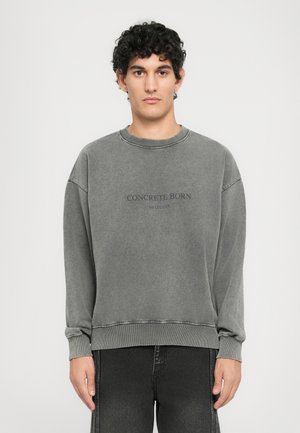 Ungdommelig mann med kort, krøllete hår iført en utydelig grå hettegenser med påskriften "CONCRETE BORN" og mørke vaskede jeans, stående mot en ensfarget bakgrunn.