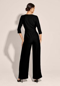 Schwarzer Jumpsuit mit Dreiviertelärmeln, tailliertem Oberteil und weiten Hosenbeinen. Verfügt über einen dezenten Reißverschluss hinten und eine glatte, matte Textur.