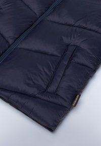 Marinblå vest med fyld, der har en frontlynlås, vandret quiltet design og en sidelomme. Blød tekstur med en sortmærke detalje forneden.