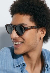 Joven con cabello rizado sonríe, usando gafas de sol de carey, un pendiente rectangular negro y una camisa de chambray azul claro.