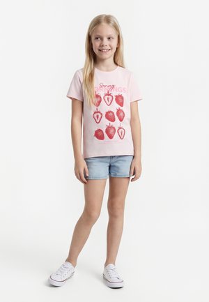 Noor tüdruk naeratab, kannab roosat t-särki punaste maasika trükkide ja tekstiga, teksapükse ja valgeid tossusid, seisab valgel taustal.