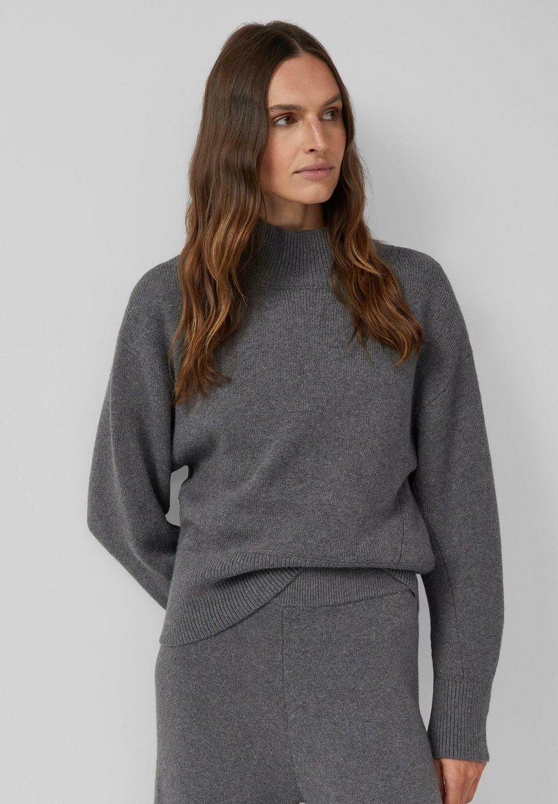 Pull gris en maille avec un col montant, des épaules tombantes et des poignets côtelés. Texture douce et coupe décontractée. Associé à un bas assorti.
