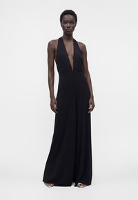 Personne grande portant une combinaison noire sans manches avec un profond décolleté en V et des jambes larges, debout devant un fond clair uni.