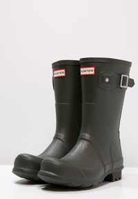 Schwarze Gummiregenstiefel mit abgerundeter Spitze, strukturiertem Grip-Sohle, verstellbaren Schnallenakzenten und rot-weißer "HUNTER"-Markierung.