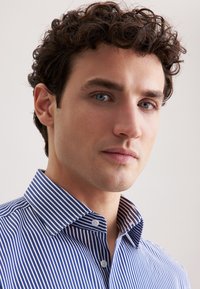Giovane con capelli ricci e occhi azzurri che indossa una camicia a righe blu e bianche con bottoni, guarda tranquillamente verso la camera.