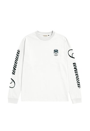 Młody mężczyzna w czerwonej czapce z niebieskim logo, białej koszuli z długim rękawem z czarnym tekstem i logo oraz w spodniach w wzór kamuflażu, stoi z rękami w kieszeniach.