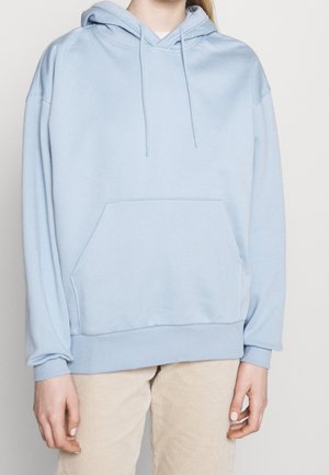 Personne portant un sweat à capuche bleu clair oversized avec poche avant et pantalon en velours côtelé de couleur crème sur fond blanc.
